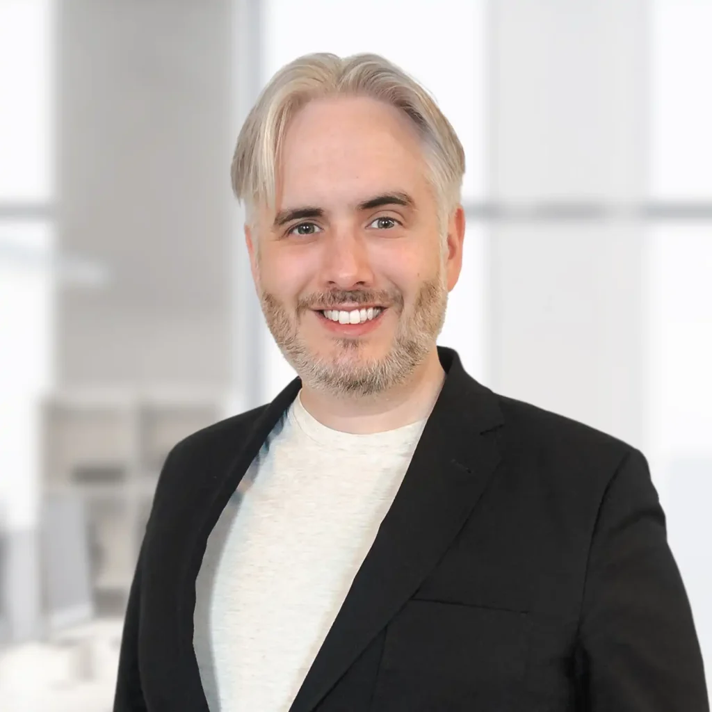 A man with light hair and a beard, wearing a black blazer and white shirt, smiles at the camera in a bright office setting.