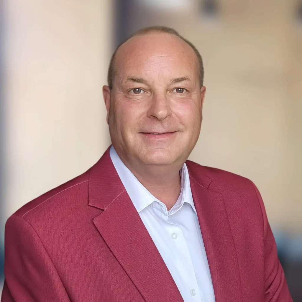 A middle-aged man wearing a red blazer and white shirt poses in front of a blurred indoor background, looking at the camera and smiling.