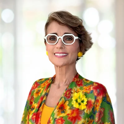 Woman wearing white glasses, yellow earrings, and a floral jacket with a yellow flower pin, standing and smiling with a blurred background.
