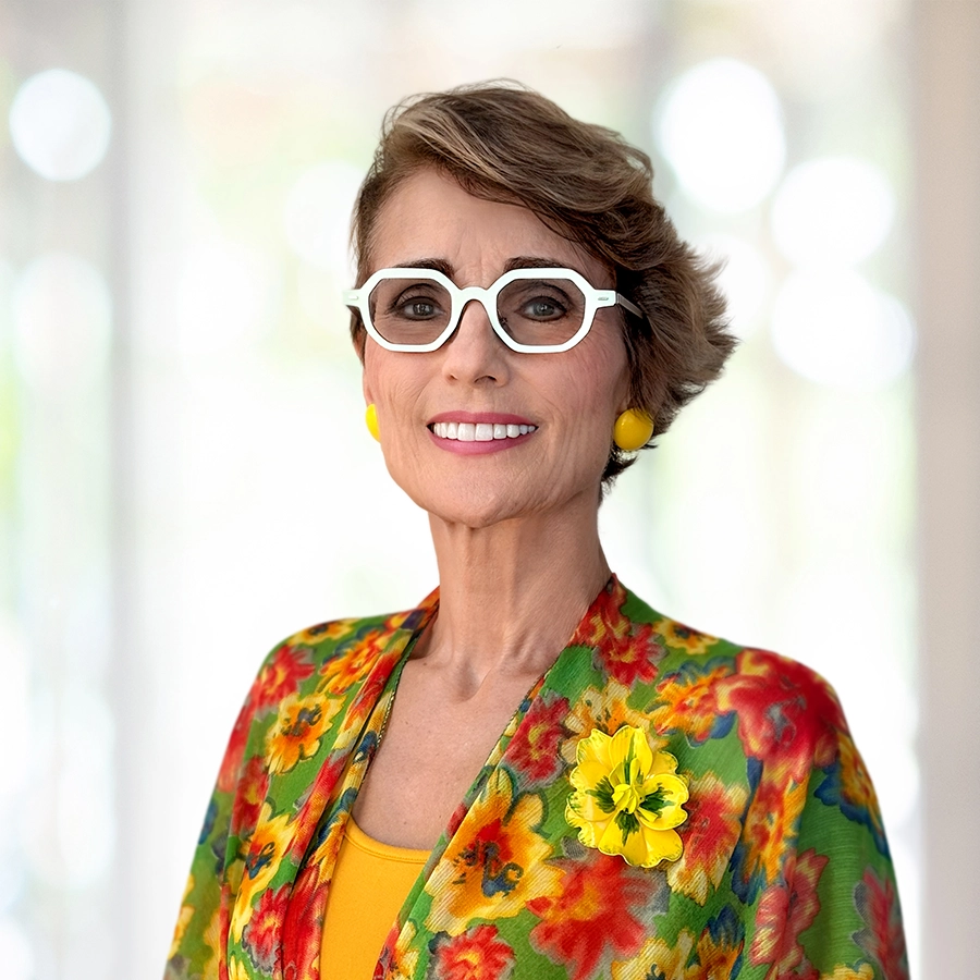 Woman wearing white glasses, yellow earrings, and a floral jacket with a yellow flower pin, standing and smiling with a blurred background.