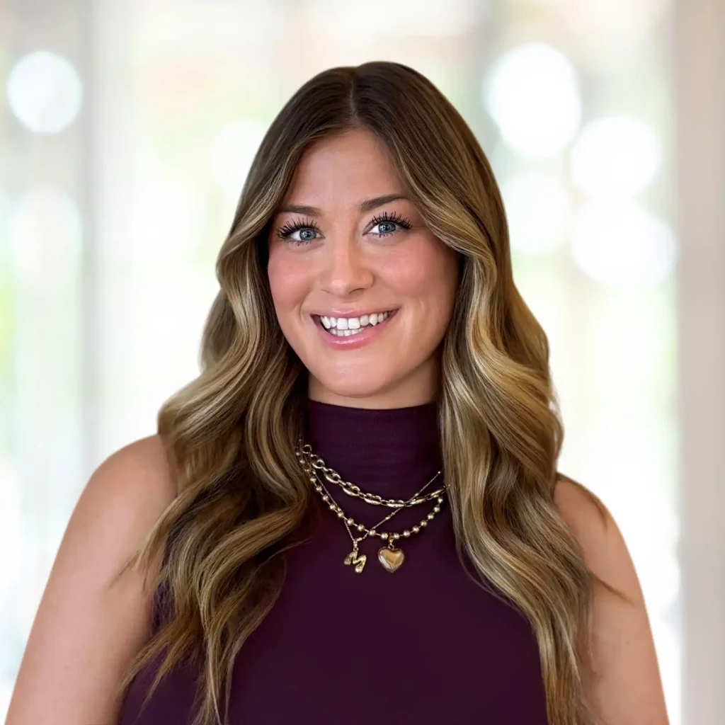A woman with long wavy brown hair, wearing a sleeveless dark purple top and layered gold necklaces, smiles at the camera in a softly lit indoor setting.