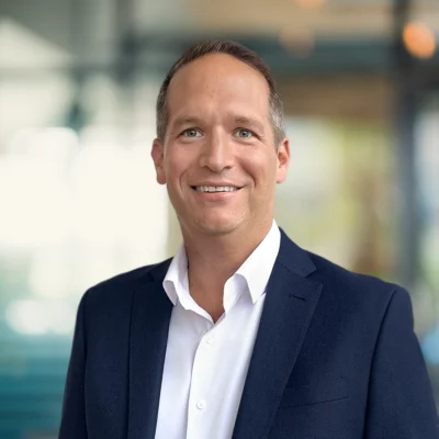 A man in a navy suit and white shirt smiles at the camera in a modern, blurred office setting.