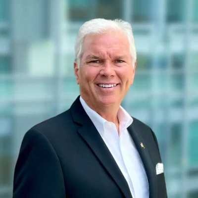 Smiling older man with white hair wearing a black suit jacket, white shirt, and pocket square, standing in front of a blurred glass building background.