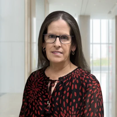 A woman with long brown hair and glasses wears a black blouse with red patterns, standing in a modern, well-lit indoor space with large windows.