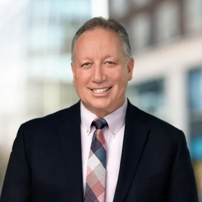 Middle-aged man wearing a suit jacket, light pink shirt, and patterned tie, smiling at the camera with a blurred cityscape background.
