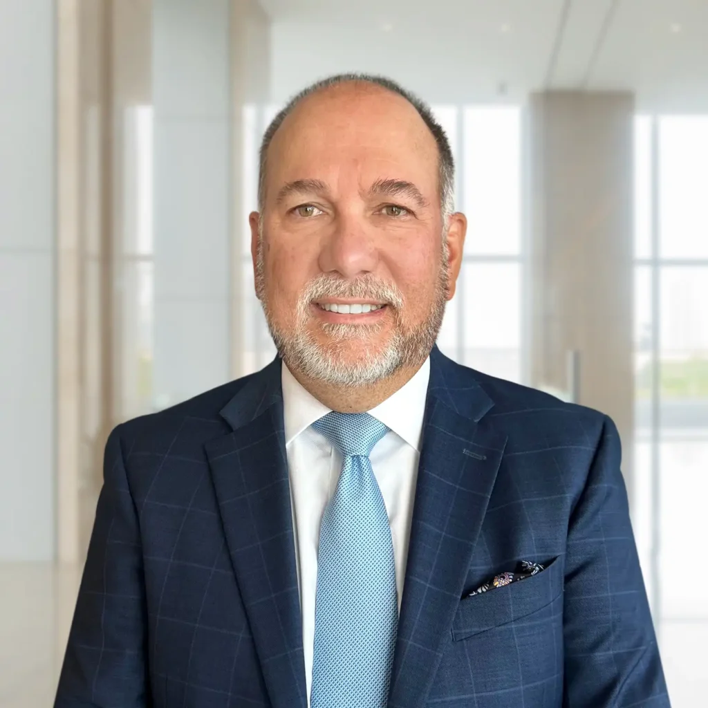 Man in a blue suit and light blue tie stands indoors in a modern, bright office setting with large windows and light-colored walls.