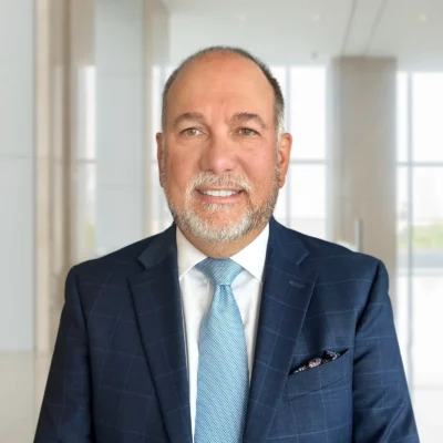 Man in a blue suit and light blue tie stands indoors in a modern, bright office setting with large windows and light-colored walls.
