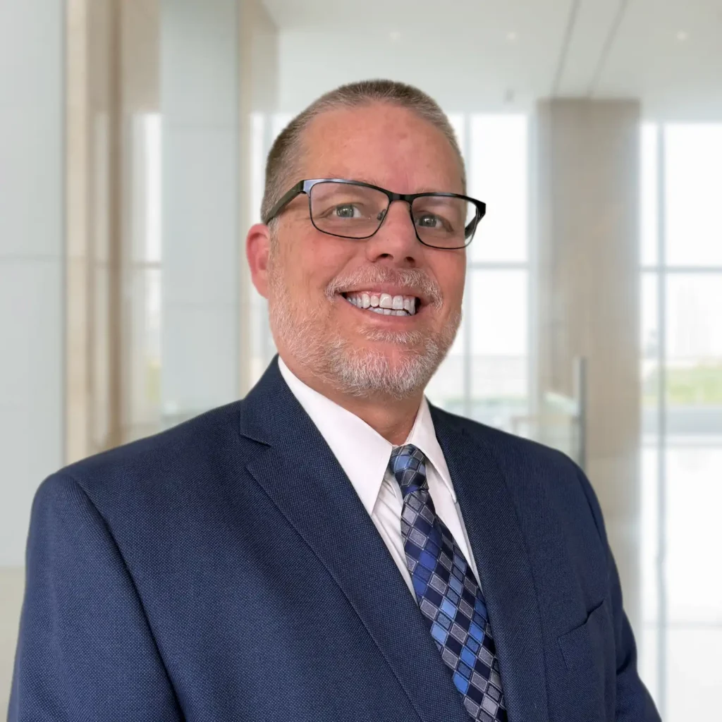 A man in a blue suit, patterned tie, and glasses smiles while standing in a modern, bright office setting with large windows.