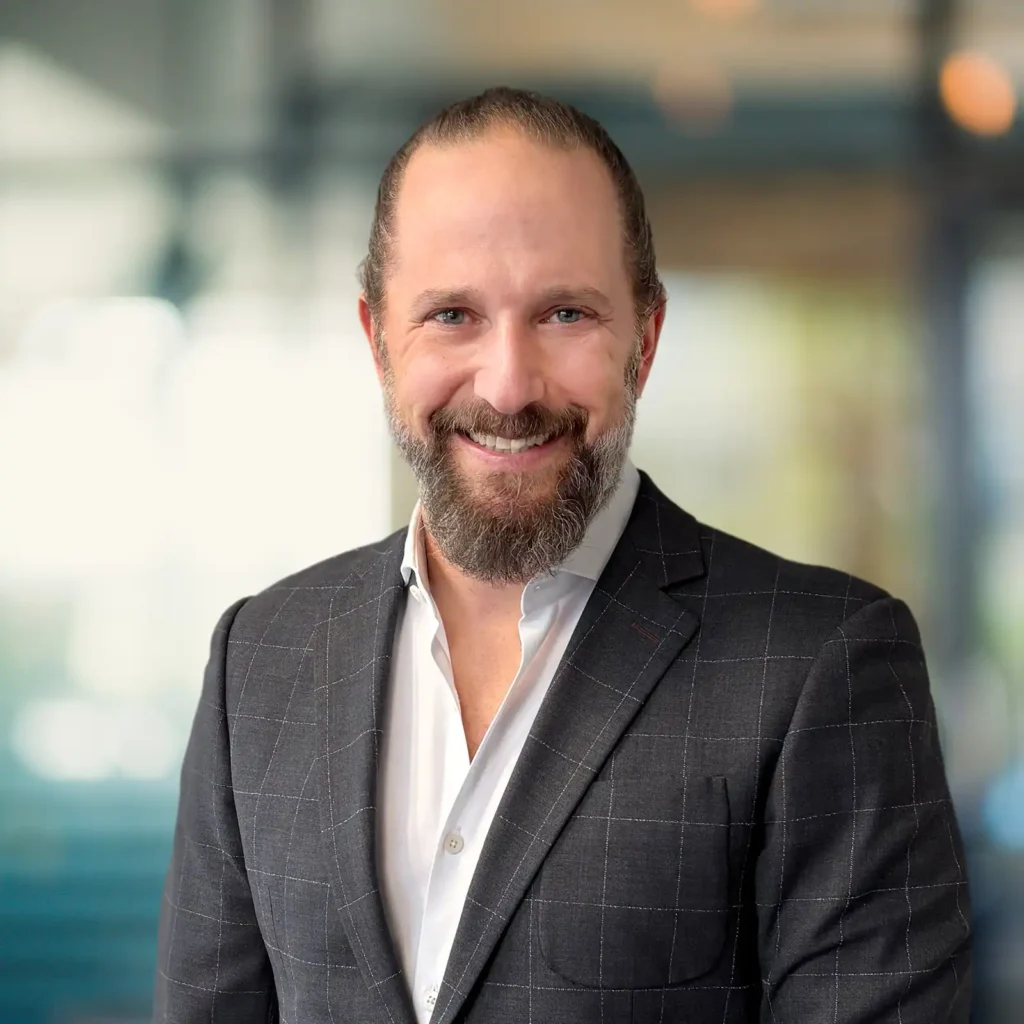 Man with a beard and long hair tied back, wearing a checked dark suit and white shirt, smiling in a modern office setting with blurred background.