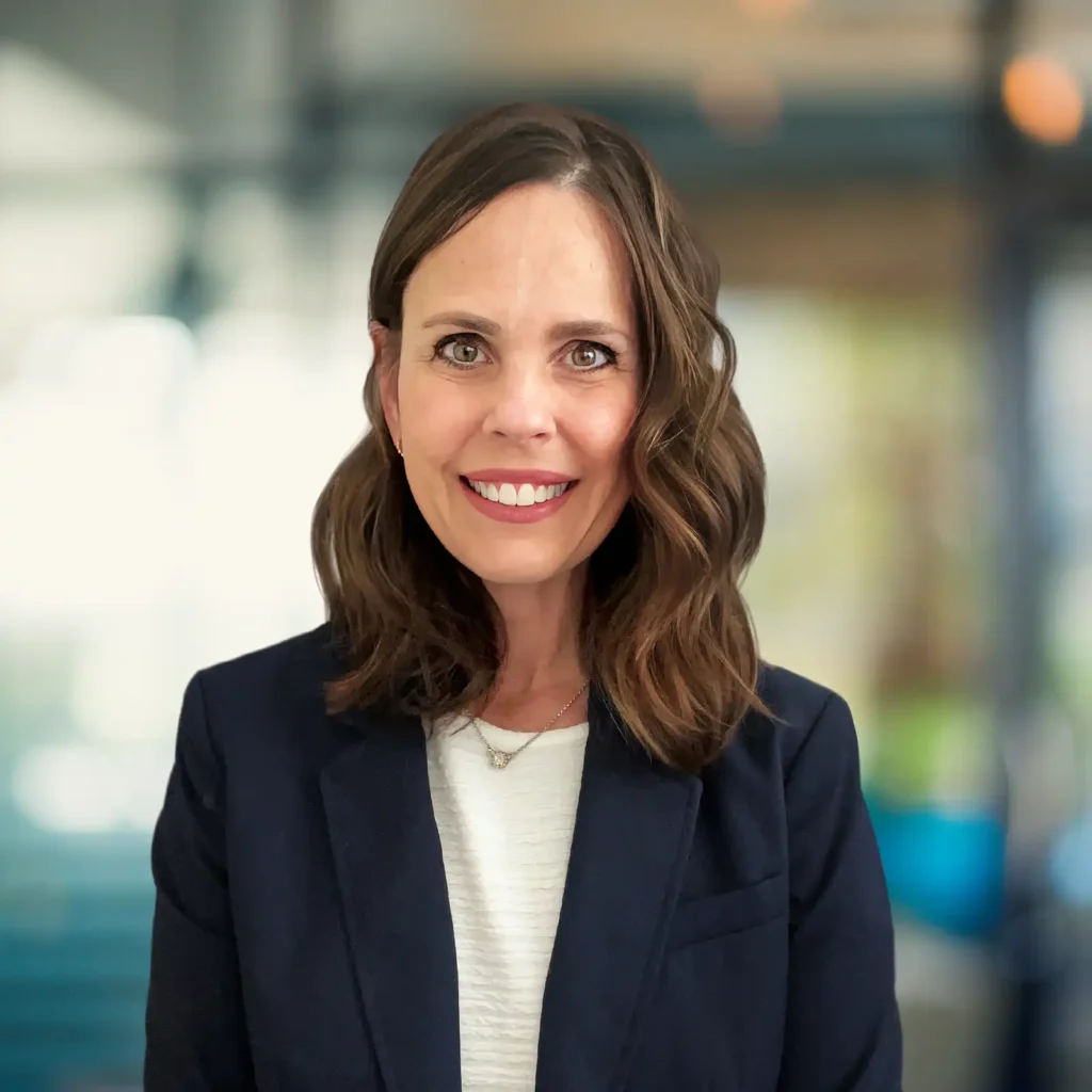 A woman with shoulder-length brown hair in loose waves wears a navy blazer and white top, smiling at the camera in a blurred office setting.