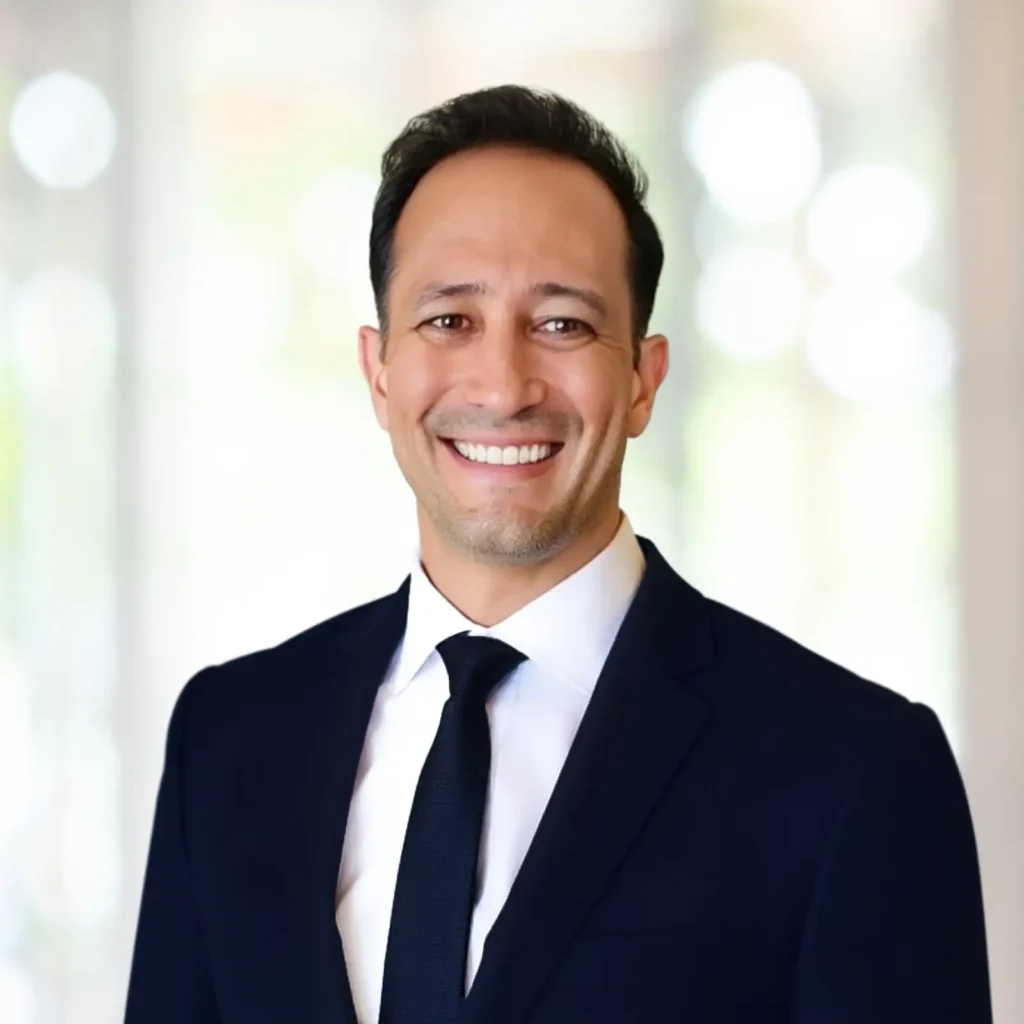 A man in a dark suit and tie smiles at the camera, standing in front of a softly blurred, light-filled background.