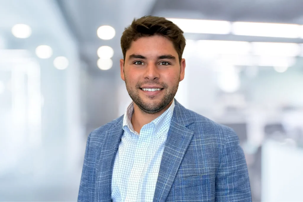 A man with short brown hair and a trimmed beard, wearing a blue plaid blazer and checked shirt, stands in a modern office with blurred lights in the background.
