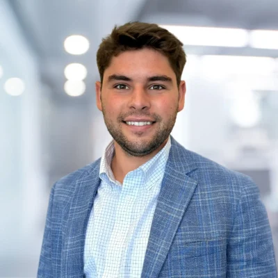 A man with short brown hair and a trimmed beard, wearing a blue plaid blazer and checked shirt, stands in a modern office with blurred lights in the background.