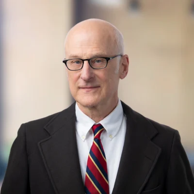 Bald man wearing glasses, a dark suit, white shirt, and a red striped tie, standing in front of a blurred background.