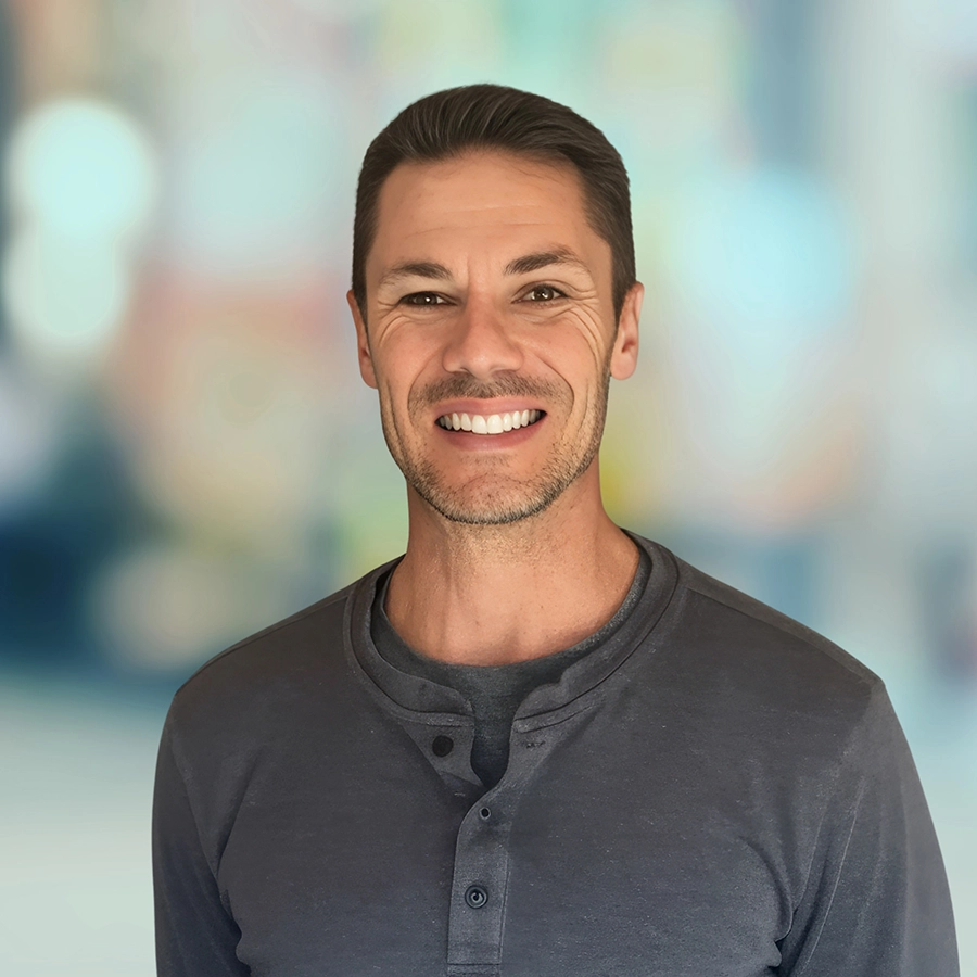 Man with short brown hair wearing a dark gray henley shirt stands smiling in front of a softly blurred, colorful background.