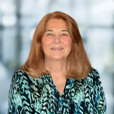 A middle-aged woman with shoulder-length light brown hair smiles at the camera, wearing a blue patterned blouse against a blurred indoor background.