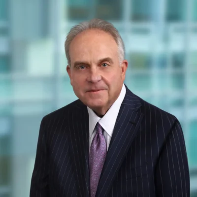An older man in a dark pinstripe suit and purple tie poses for a formal portrait against a blurred office background.