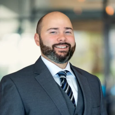 A man with a beard and mustache, wearing a gray suit, white shirt, and striped tie, smiles at the camera in a blurred office setting.
