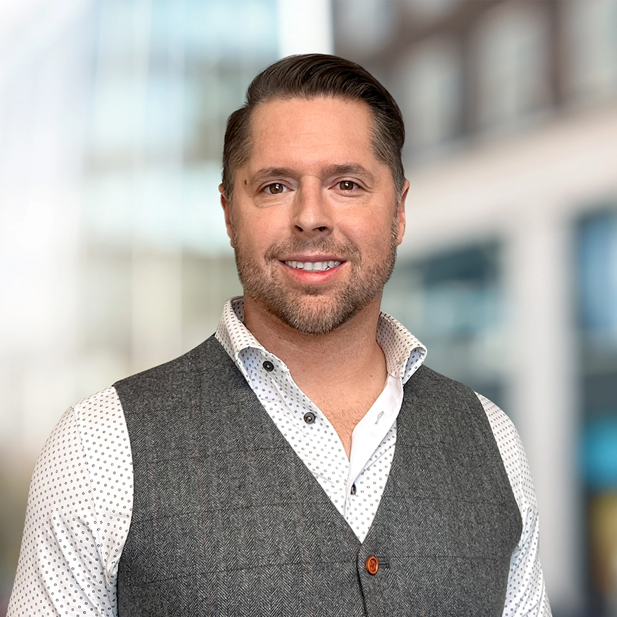 A man with short brown hair and a trimmed beard is wearing a white patterned shirt and gray vest, standing in front of a blurred building background.