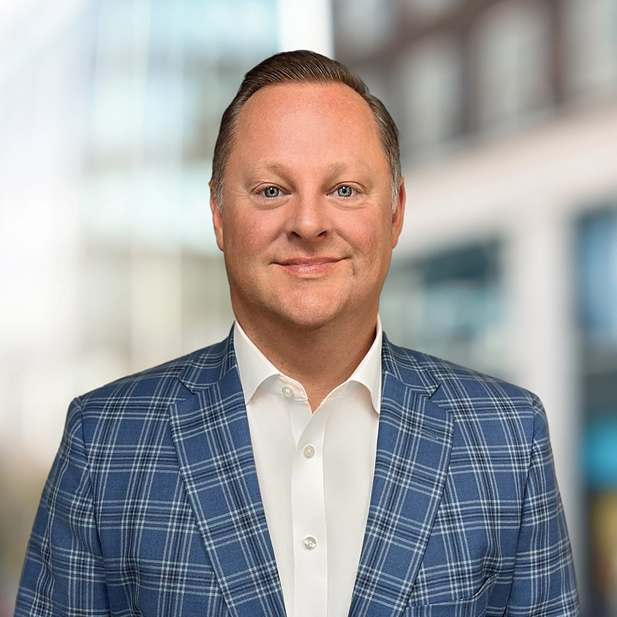 A man in a blue plaid suit jacket and white shirt poses in front of a blurred urban background.