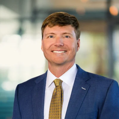 A man in a blue suit, white shirt, and patterned yellow tie smiles at the camera against a blurred office background.