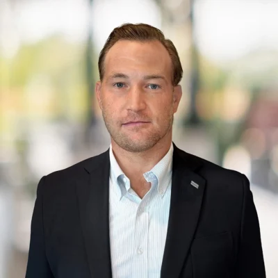 A man in a dark suit jacket and light blue shirt stands indoors against a blurred background, facing the camera with a neutral expression.