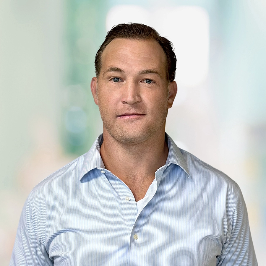 A man with short brown hair wearing a light blue button-up shirt stands in front of a blurred indoor background.