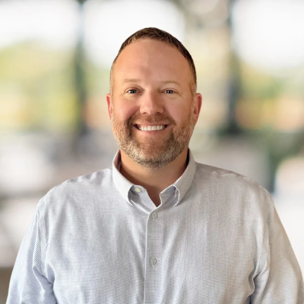 A man with short hair and a beard smiles at the camera. He is wearing a light-colored button-up shirt and stands in front of a blurred background.