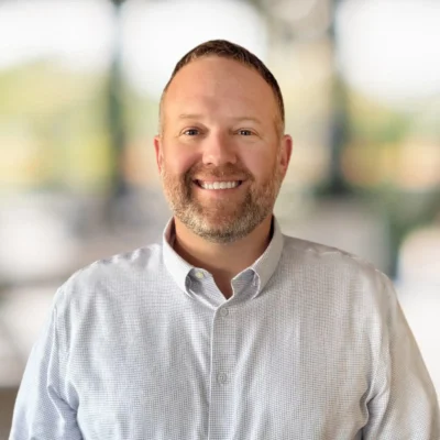 A man with short hair and a beard smiles at the camera. He is wearing a light-colored button-up shirt and stands in front of a blurred background.