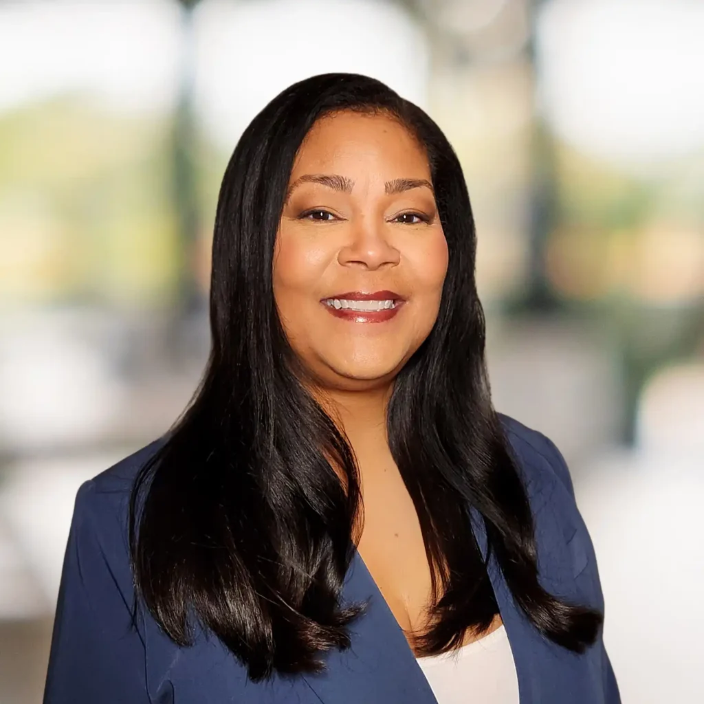 A woman with long black hair, wearing a blue blazer and white top, smiles at the camera with a blurred background.