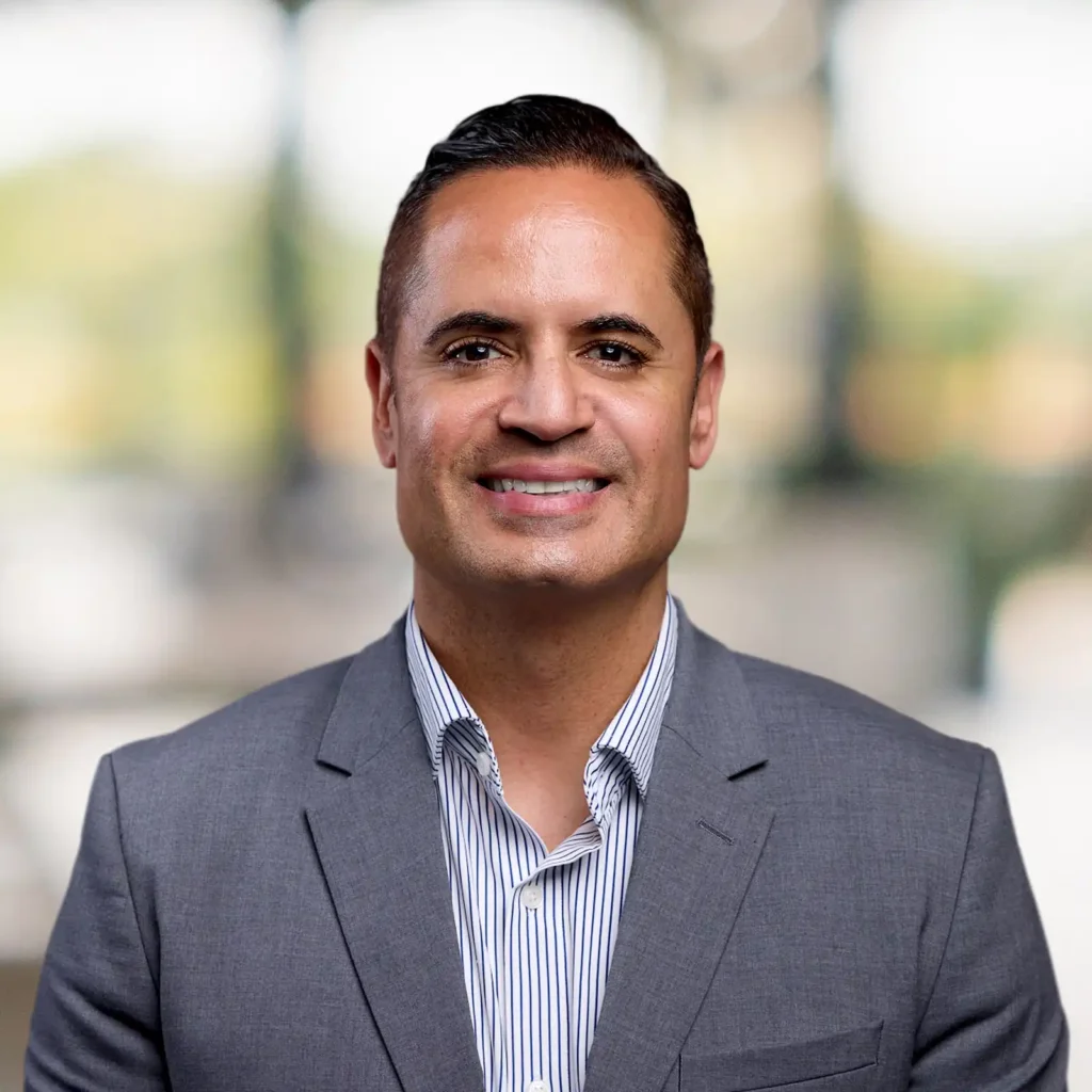A man wearing a grey suit and striped shirt smiles at the camera, standing in front of a blurred indoor background.