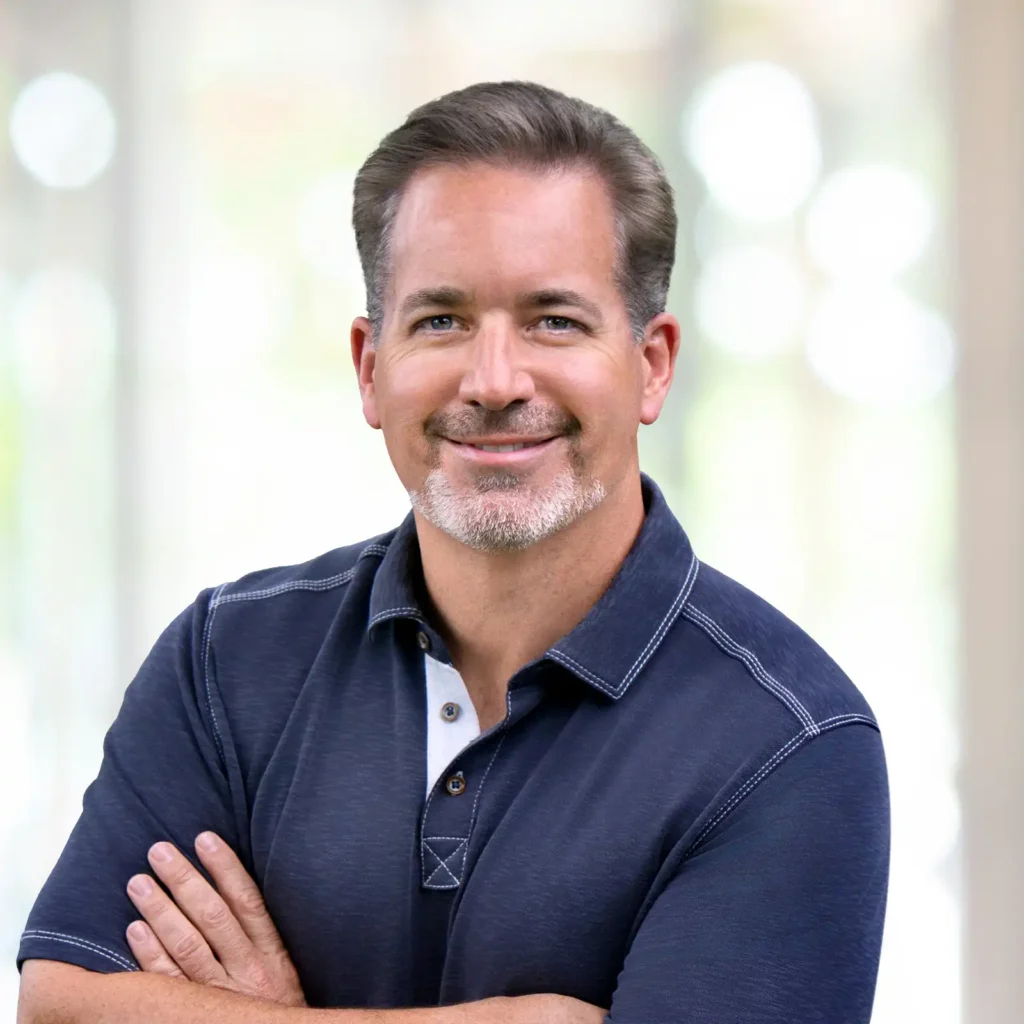 A man with short brown hair and a trimmed beard, wearing a navy blue polo shirt, stands with arms crossed and smiles at the camera in front of a blurred light background.