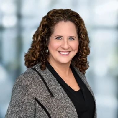 A woman with curly brown hair, wearing a gray blazer over a black top, smiles at the camera against a blurred blue and white background.
