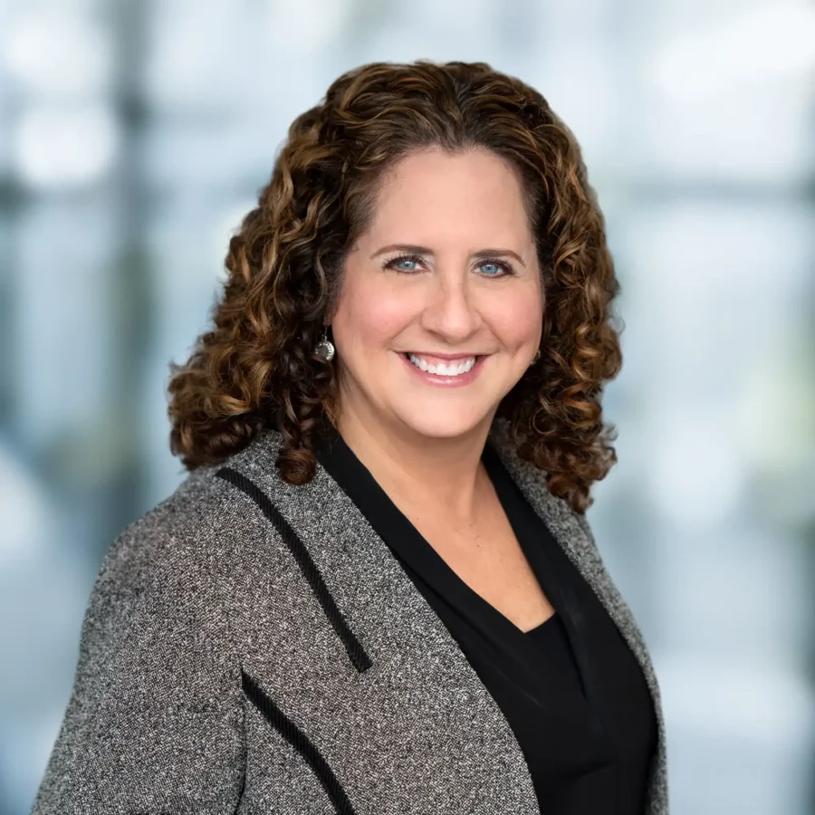 A woman with curly brown hair, wearing a gray blazer over a black top, smiles at the camera against a blurred blue and white background.