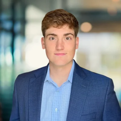 A young man with short brown hair in a blue suit jacket and light blue shirt stands in front of a blurred indoor background.