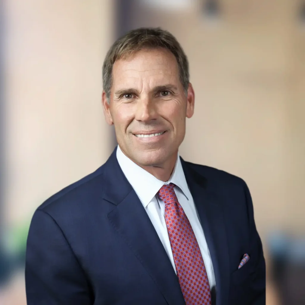 A man in a navy blue suit, white shirt, and red patterned tie is smiling at the camera against a blurred beige background.