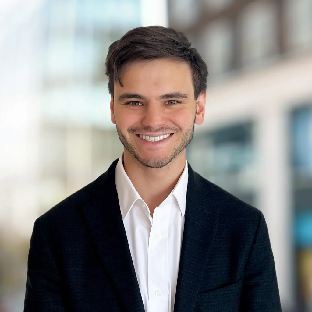 A young man in a dark blazer and white shirt smiles at the camera with a blurred office building background.