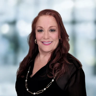 Smiling woman with long, reddish-brown hair wearing a black blouse and a long necklace, posed in front of a blurred indoor background.