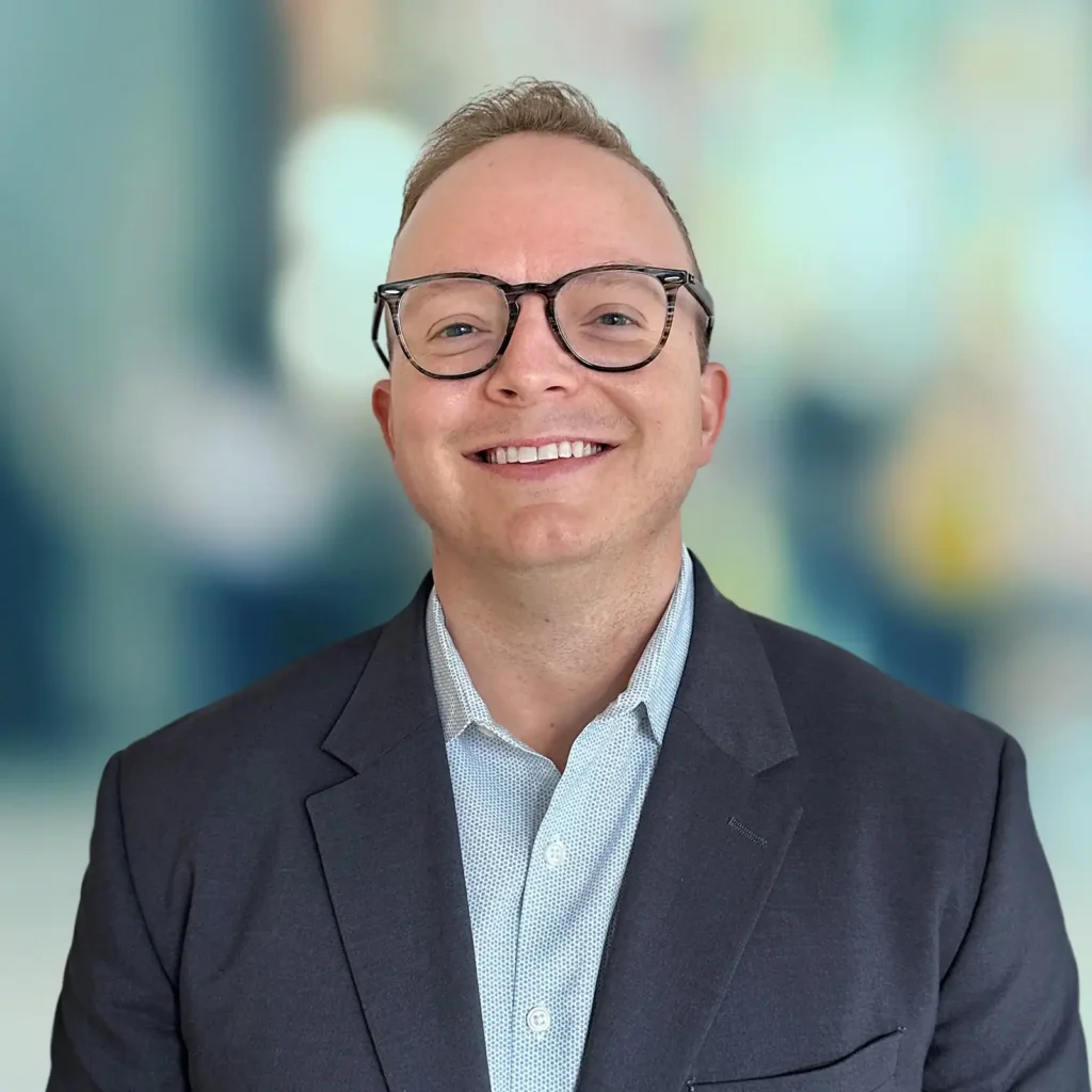 A person with short light hair and glasses smiles at the camera, wearing a dark blazer and light-colored shirt, with a blurred background.