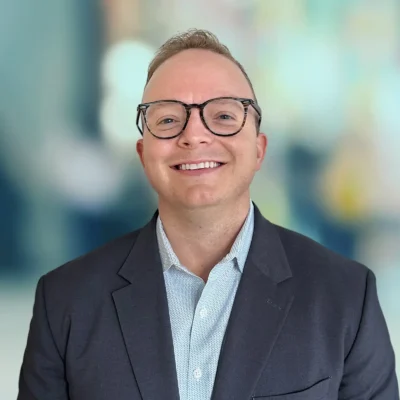 A person with short light hair and glasses smiles at the camera, wearing a dark blazer and light-colored shirt, with a blurred background.