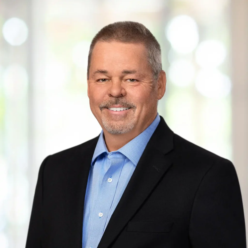 A middle-aged man with short hair and a goatee, wearing a blue shirt and dark blazer, smiles in front of a softly blurred light background.