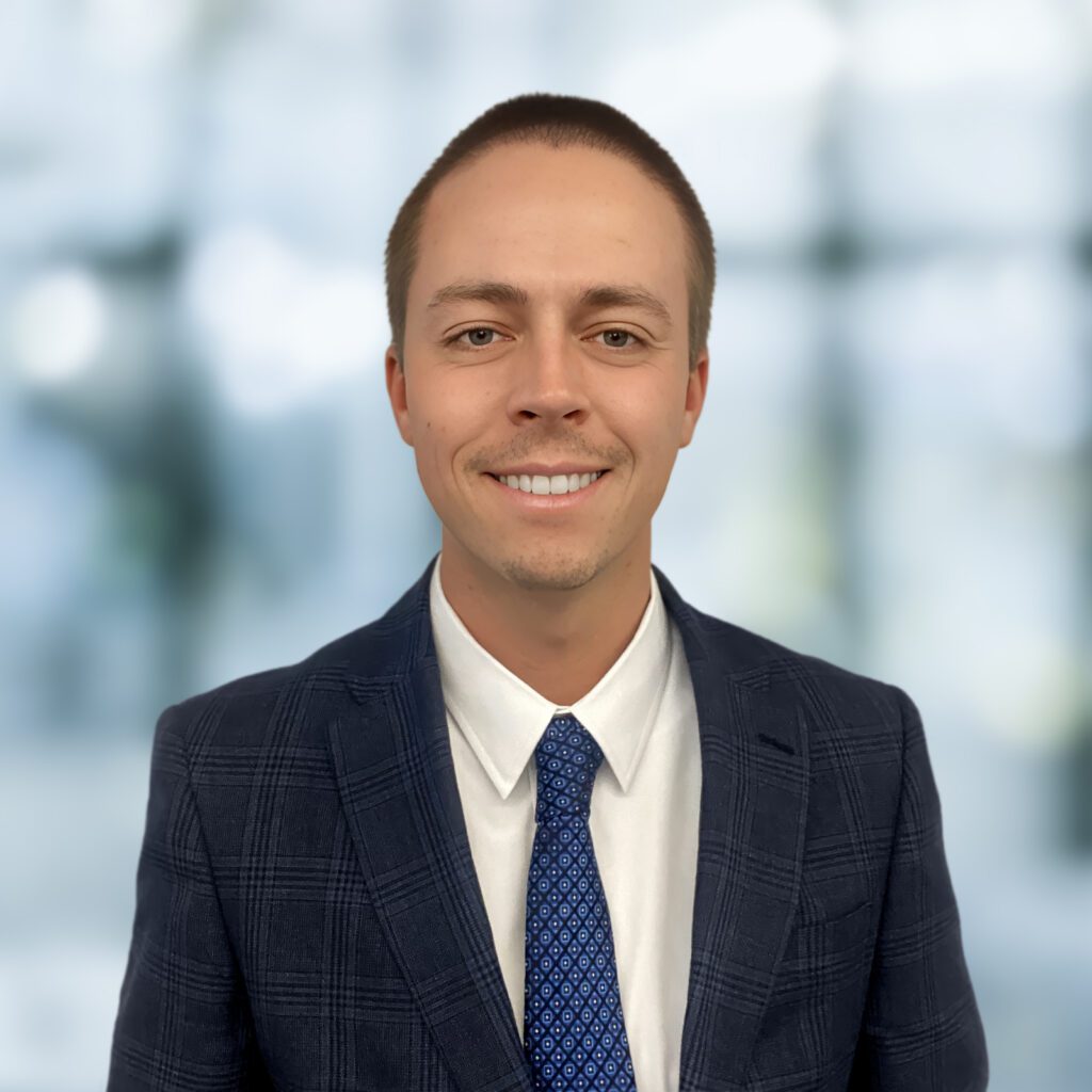 A man wearing a dark plaid suit, white shirt, and blue patterned tie stands in front of a blurred indoor background, smiling at the camera.
