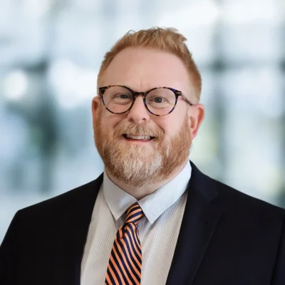 A man with short red hair, a beard, and glasses is wearing a dark suit, light shirt, and striped tie, standing in front of a blurred background.