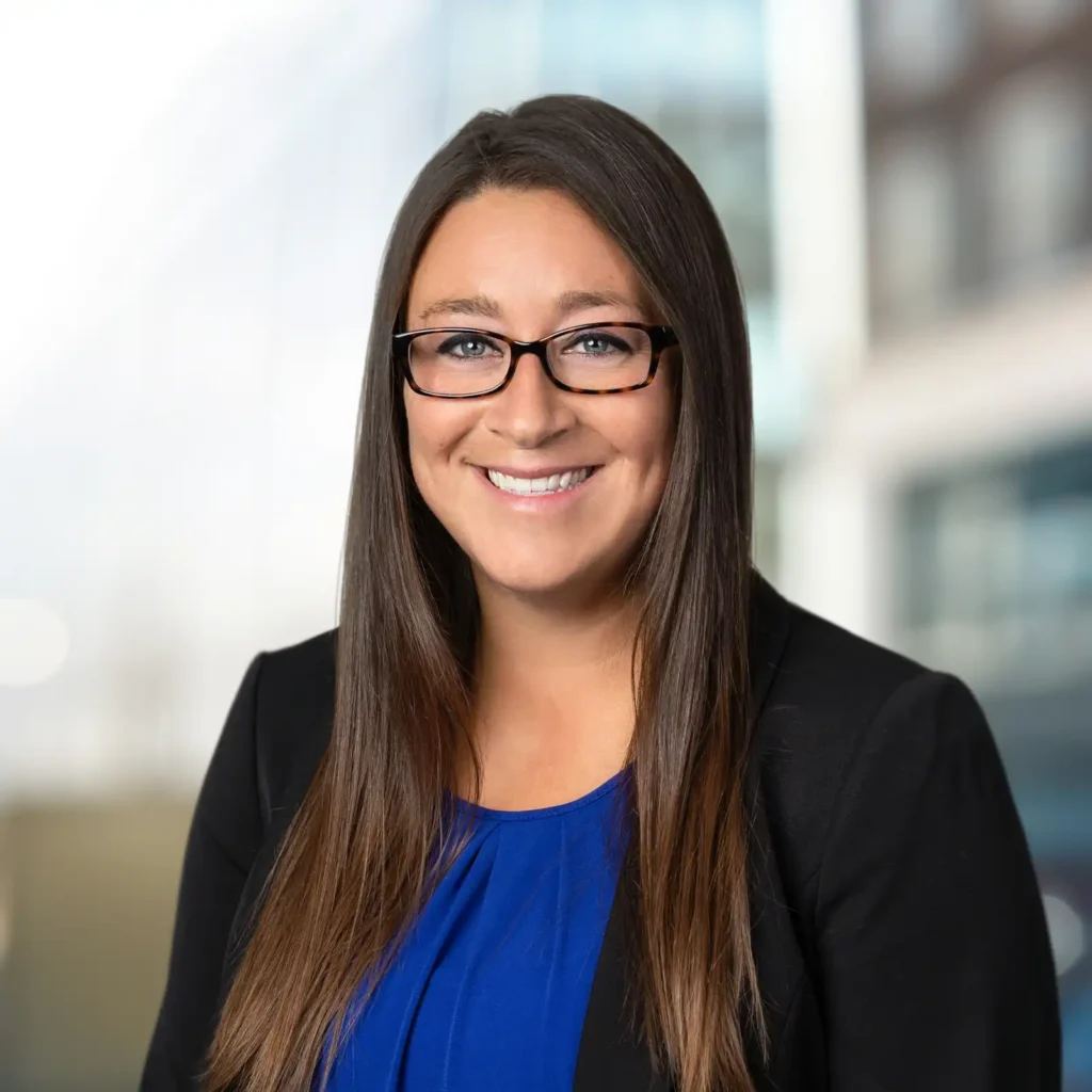 Woman with long brown hair and glasses, wearing a blue blouse and black blazer, smiling in front of a blurred office background.