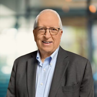 Older man with short gray hair, glasses, and a smile, wearing a light blue shirt and dark blazer, standing in a brightly lit office setting.