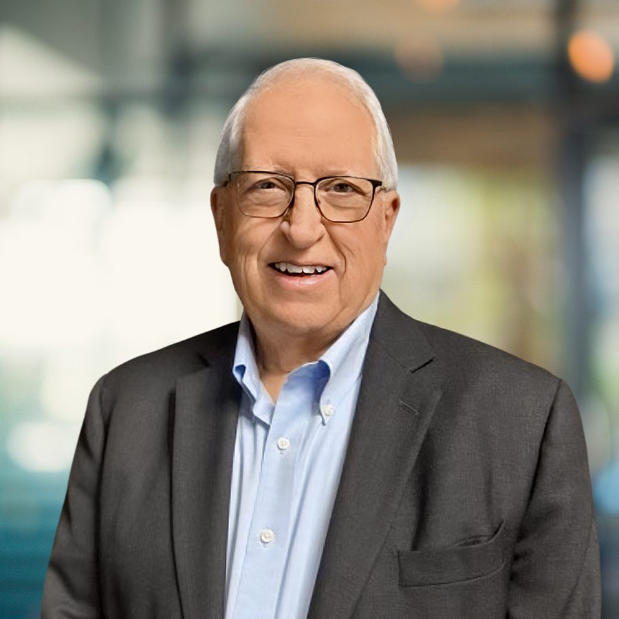 Older man with short gray hair, glasses, and a smile, wearing a light blue shirt and dark blazer, standing in a brightly lit office setting.