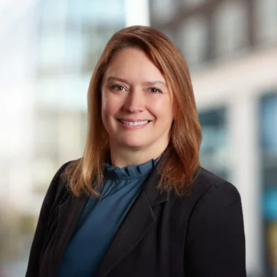 A woman with straight, light brown hair is smiling, wearing a dark blazer and blue blouse, standing in front of a blurred office building background.