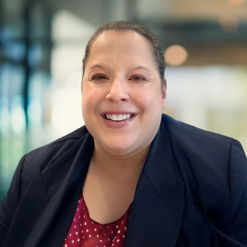 A woman with dark hair pulled back, wearing a navy blazer over a red polka-dot blouse, smiles at the camera in a brightly lit indoor setting.
