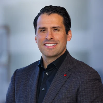 Man with dark hair wearing a dark checkered blazer and black shirt, smiling at the camera against a blurred background.
