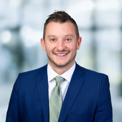 A man wearing a blue pinstripe suit, light blue shirt, and green striped tie smiles at the camera with a blurred background.